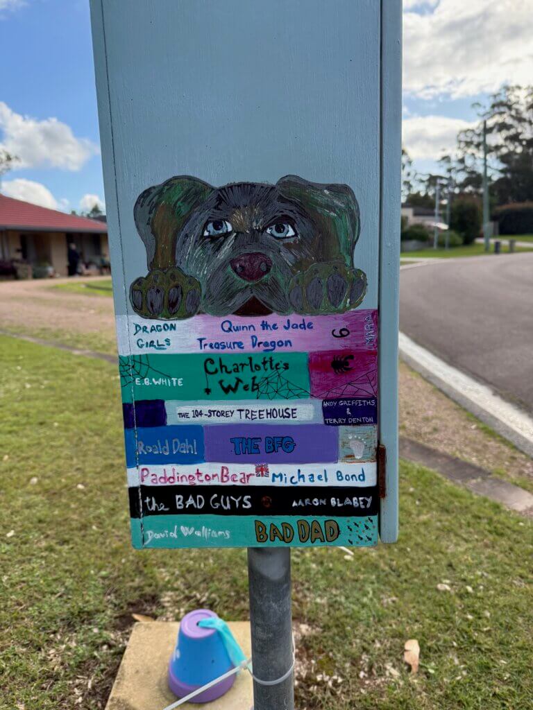 Side of street library with painting of book spines and a dog looking over them.