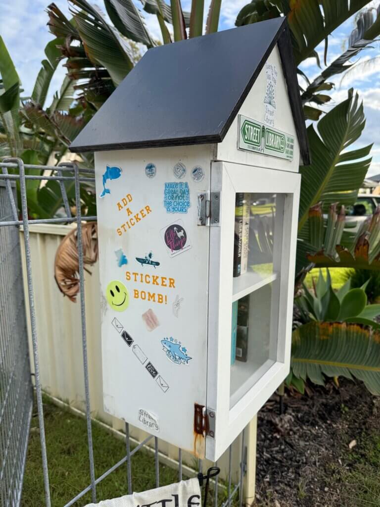 Side of a street library covered in stickers, with instructions to "ADD A STICKER' & 'STICKER BOMB'!