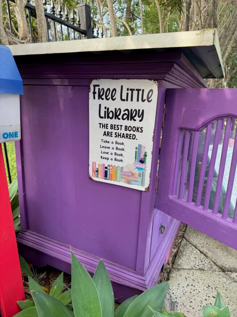 A street library made out of an old tv cupboard painted purple, with the door open and a sign saying "Free Little Library. The Best Books are Shared. Take a Book. Leave a Book. Love a Book. Keep a Book."
