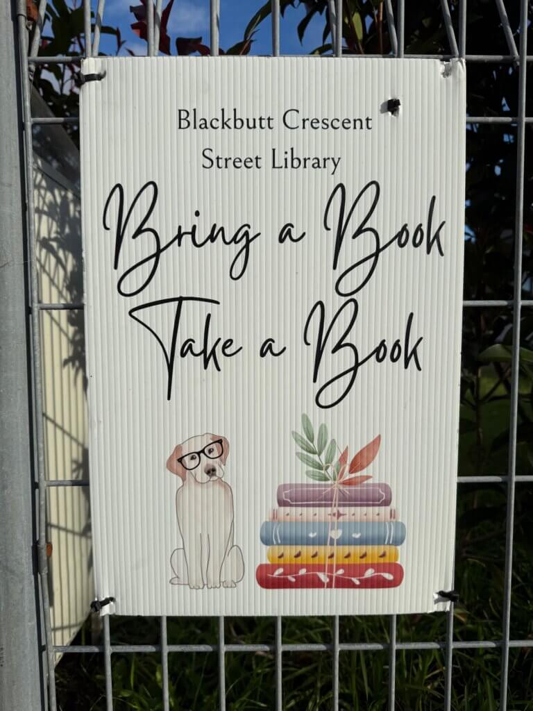 Sign on a fence saying "Blackbutt Crescent Street Library. Bring a Book. Take a Book." above a drawing of a dog wearing glasses and a pile of books topped with some leaves.